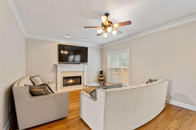 living room with light wood finished floors, crown molding, ceiling fan, baseboards, and a warm lit fireplace