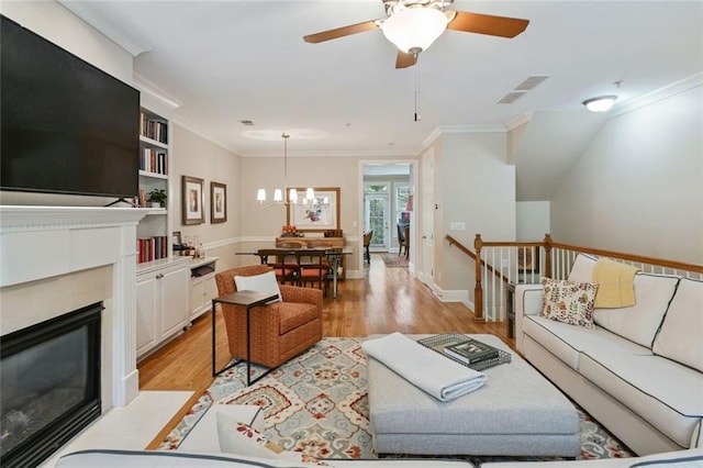 living room with visible vents, a fireplace with flush hearth, crown molding, ceiling fan with notable chandelier, and light wood-style flooring