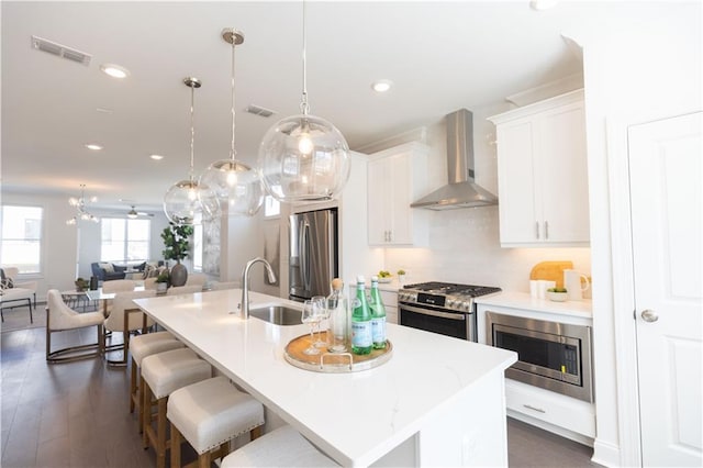 kitchen featuring wall chimney exhaust hood, stainless steel appliances, sink, pendant lighting, and an island with sink