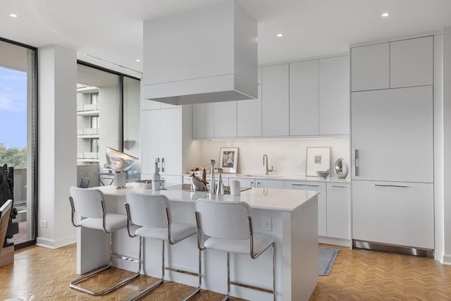 kitchen with white cabinetry, an island with sink, sink, wall chimney range hood, and light parquet floors