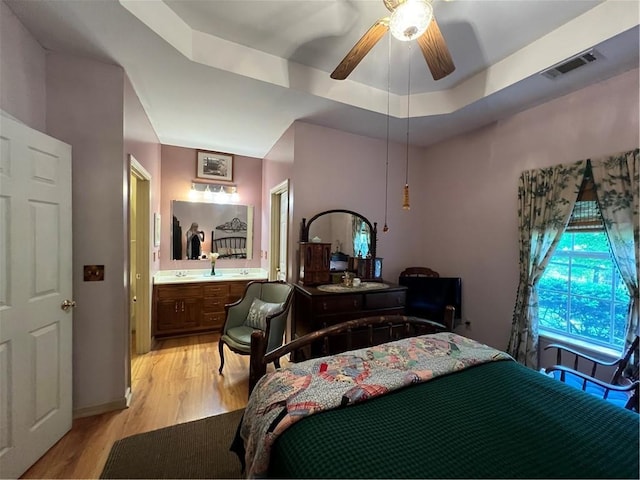 bedroom with light wood-style flooring, ensuite bathroom, a raised ceiling, and a ceiling fan