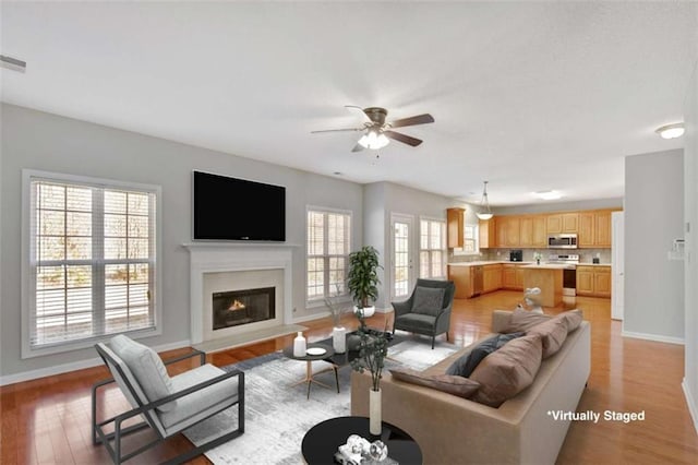 living area featuring light wood-style flooring, baseboards, a ceiling fan, and a glass covered fireplace