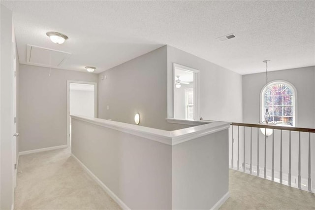 hallway with a textured ceiling, light colored carpet, visible vents, baseboards, and attic access