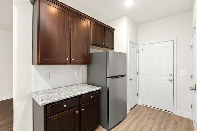 kitchen with backsplash, light wood-type flooring, dark brown cabinets, light stone counters, and freestanding refrigerator