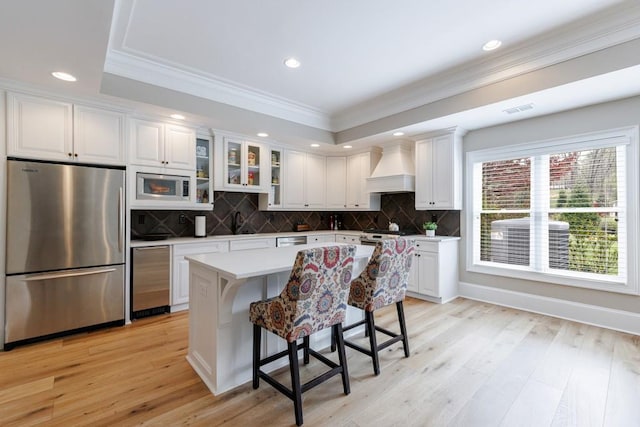 kitchen with light wood-type flooring, custom range hood, backsplash, white cabinets, and appliances with stainless steel finishes