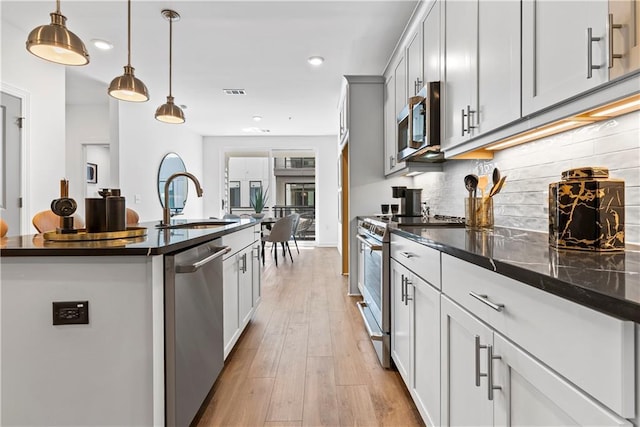 kitchen with stainless steel appliances, light hardwood / wood-style flooring, backsplash, decorative light fixtures, and a center island with sink