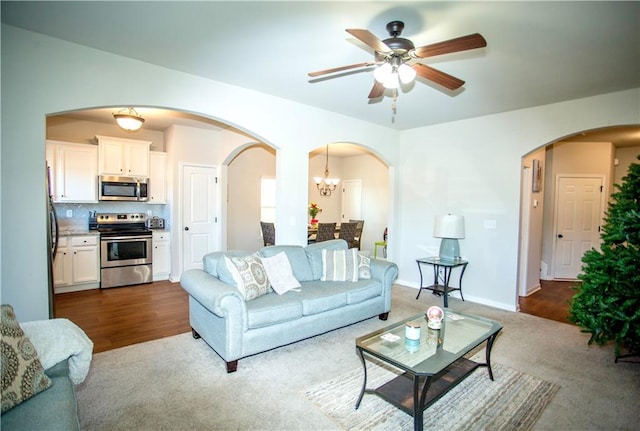 living room with arched walkways, ceiling fan with notable chandelier, dark carpet, and baseboards