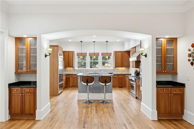 kitchen with glass insert cabinets, stainless steel range with gas cooktop, light wood-type flooring, brown cabinetry, and wall chimney exhaust hood