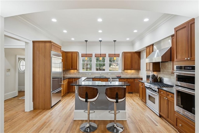 kitchen with a center island, wall chimney range hood, high quality appliances, ornamental molding, and brown cabinets