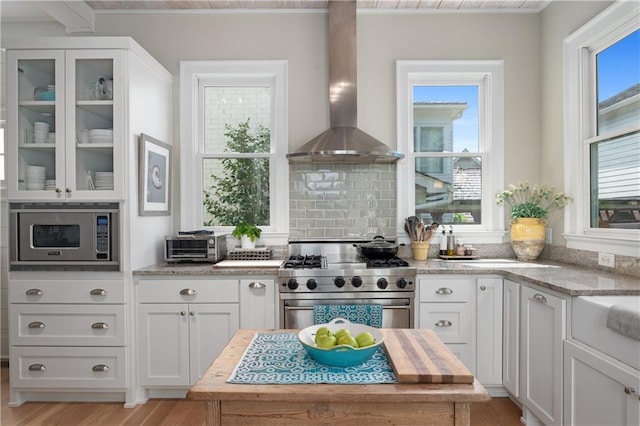 kitchen with white cabinets, appliances with stainless steel finishes, light wood finished floors, wall chimney exhaust hood, and glass insert cabinets