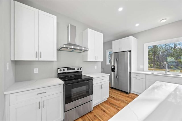 kitchen with a sink, light wood-style floors, white cabinets, appliances with stainless steel finishes, and wall chimney range hood