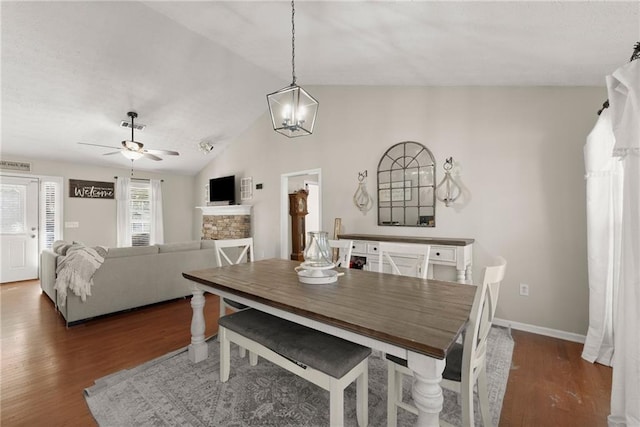 dining room featuring ceiling fan with notable chandelier, a stone fireplace, wood finished floors, and vaulted ceiling