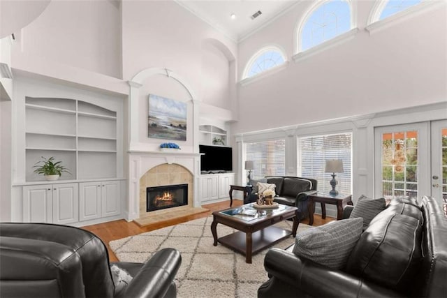 living area featuring built in features, a tiled fireplace, a towering ceiling, ornamental molding, and light wood-type flooring