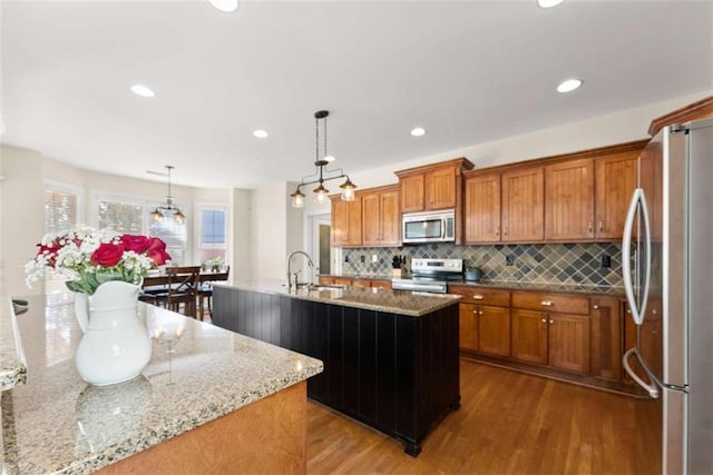 kitchen featuring a center island with sink, brown cabinetry, appliances with stainless steel finishes, pendant lighting, and a sink