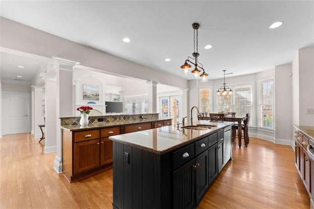 kitchen featuring a center island with sink, hanging light fixtures, stainless steel dishwasher, a sink, and ornate columns