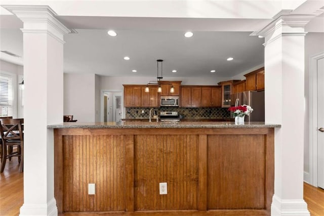 kitchen with decorative columns, appliances with stainless steel finishes, brown cabinetry, glass insert cabinets, and a peninsula