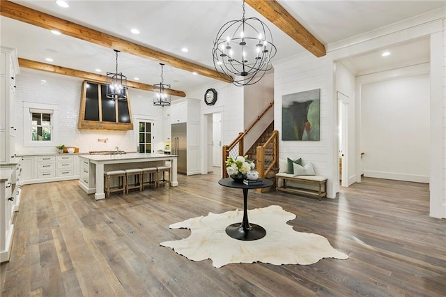 interior space featuring beamed ceiling, hanging light fixtures, a center island with sink, hardwood / wood-style floors, and white cabinetry