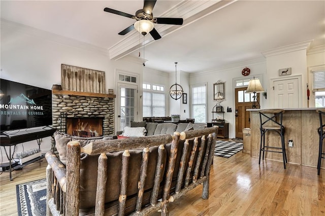 living room with ceiling fan with notable chandelier, light hardwood / wood-style flooring, crown molding, and a stone fireplace