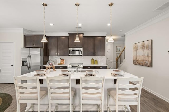 kitchen with decorative light fixtures, a kitchen island with sink, backsplash, appliances with stainless steel finishes, and dark brown cabinets