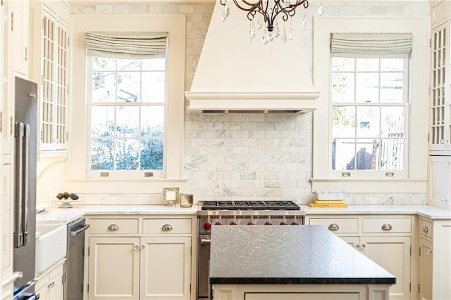 kitchen with light stone counters, custom exhaust hood, stainless steel stove, a notable chandelier, and tasteful backsplash