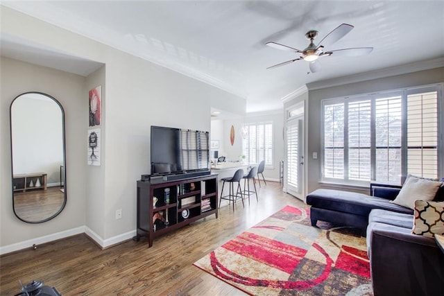 living room featuring a ceiling fan, crown molding, baseboards, and wood finished floors