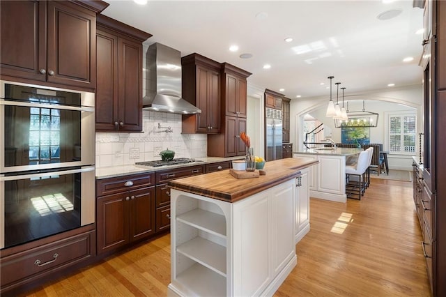 kitchen with arched walkways, appliances with stainless steel finishes, a kitchen island with sink, wall chimney range hood, and open shelves