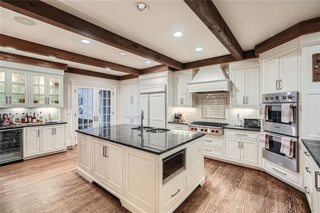 kitchen with custom exhaust hood, built in appliances, a center island with sink, dark stone countertops, and wine cooler