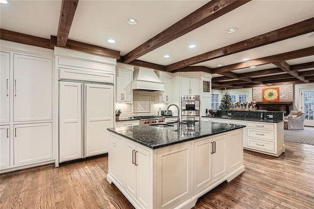 kitchen featuring premium range hood, sink, dark stone counters, and a center island with sink