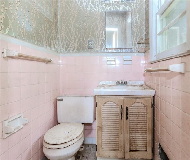 bathroom featuring tile walls, toilet, wallpapered walls, vanity, and a wainscoted wall