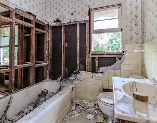 bathroom featuring wallpapered walls, tile walls, toilet, a tub to relax in, and a sink