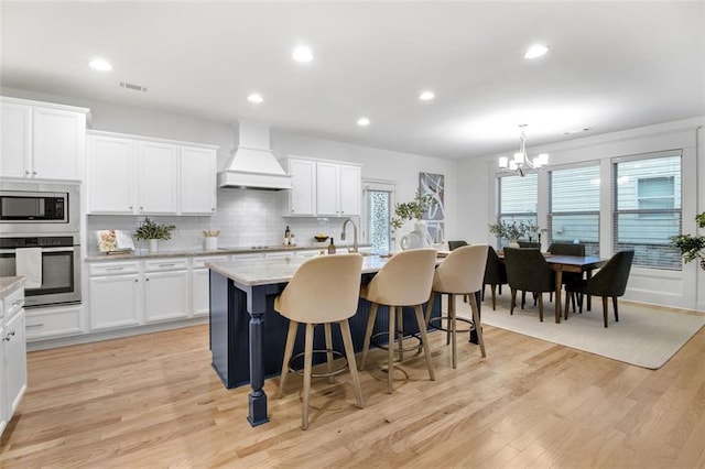 kitchen featuring premium range hood, an island with sink, a notable chandelier, white cabinetry, and appliances with stainless steel finishes