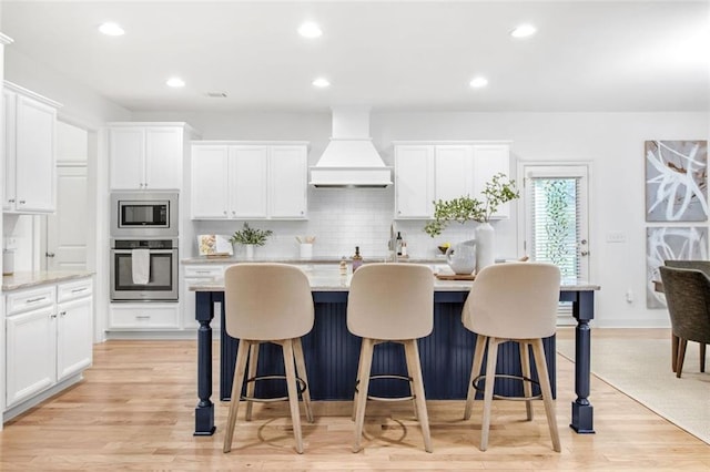 kitchen with white cabinetry, light wood-type flooring, decorative backsplash, premium range hood, and appliances with stainless steel finishes