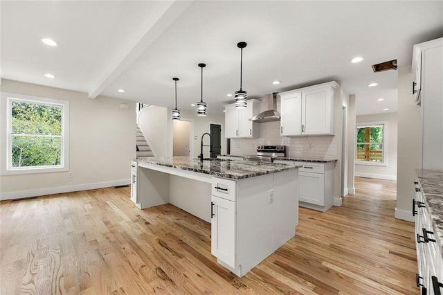 kitchen with white cabinetry, dark stone counters, light wood-type flooring, a center island with sink, and wall chimney range hood