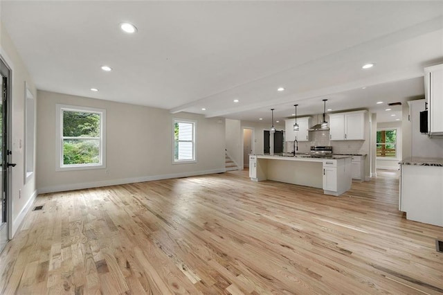 kitchen featuring light wood-type flooring, white cabinets, wall chimney exhaust hood, a center island with sink, and decorative light fixtures