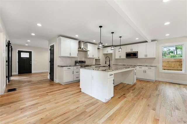 kitchen with appliances with stainless steel finishes, pendant lighting, light wood-type flooring, a kitchen island with sink, and wall chimney range hood