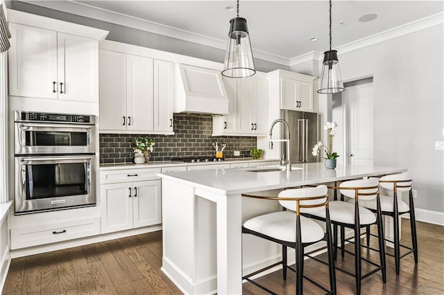 kitchen with light countertops, custom range hood, appliances with stainless steel finishes, a sink, and an island with sink