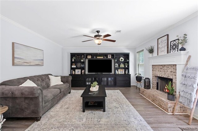 living room with a ceiling fan, visible vents, a brick fireplace, wood finished floors, and ornamental molding