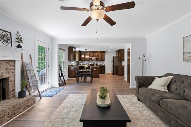 living room featuring ceiling fan, a brick fireplace, baseboards, ornamental molding, and light wood-style flooring