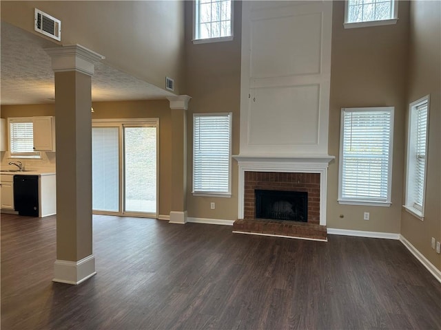 unfurnished living room with dark wood-style floors, visible vents, a brick fireplace, and decorative columns