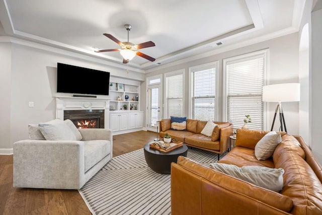 living area featuring a raised ceiling, wood finished floors, a warm lit fireplace, and ceiling fan