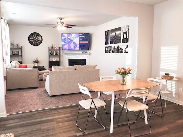 dining space with dark wood-type flooring, a fireplace, a ceiling fan, and baseboards