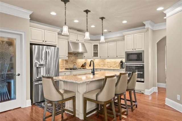 kitchen featuring pendant lighting, appliances with stainless steel finishes, a kitchen island with sink, light stone countertops, and ornamental molding