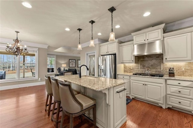 kitchen with a kitchen island with sink, stainless steel refrigerator with ice dispenser, light stone counters, ornamental molding, and decorative light fixtures