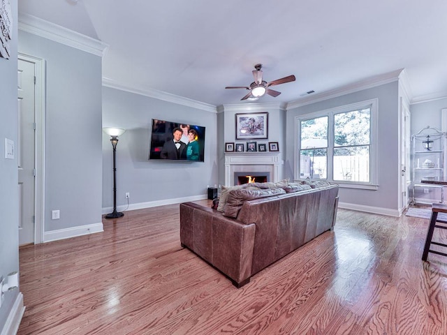 living area with a ceiling fan, wood finished floors, crown molding, baseboards, and a warm lit fireplace
