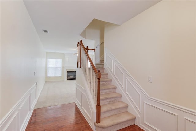 staircase featuring visible vents, a decorative wall, wainscoting, wood finished floors, and a glass covered fireplace