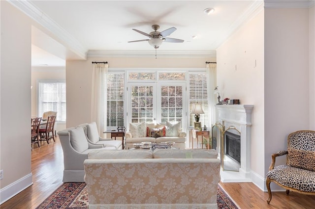living room featuring ceiling fan, wood finished floors, crown molding, baseboards, and a fireplace with raised hearth