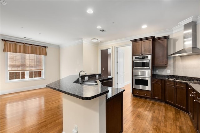 kitchen with an island with sink, appliances with stainless steel finishes, sink, and wall chimney range hood