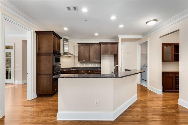 kitchen featuring stainless steel oven, dark stone counters, an island with sink, and wall chimney range hood