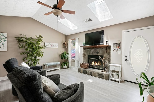 living room with a stone fireplace, light hardwood / wood-style flooring, vaulted ceiling with skylight, ceiling fan, and a textured ceiling