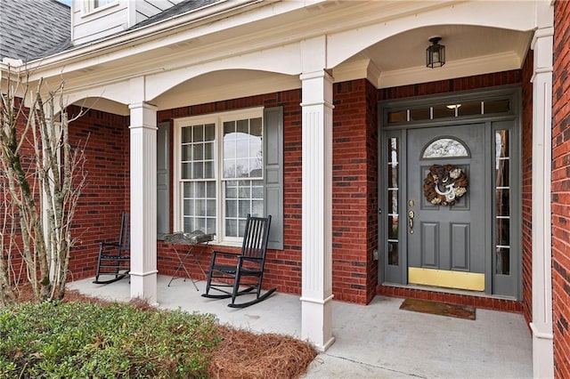 doorway to property with covered porch, brick siding, and roof with shingles